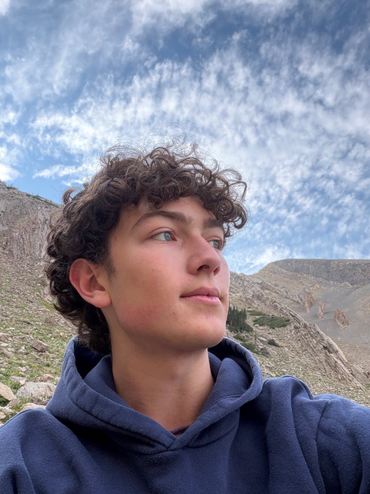 Cruce Saunders in a navy hoodie looking off to the side, curly hair against streaky white clouds and blue sky, a rocky Montana mountain slope behind him.