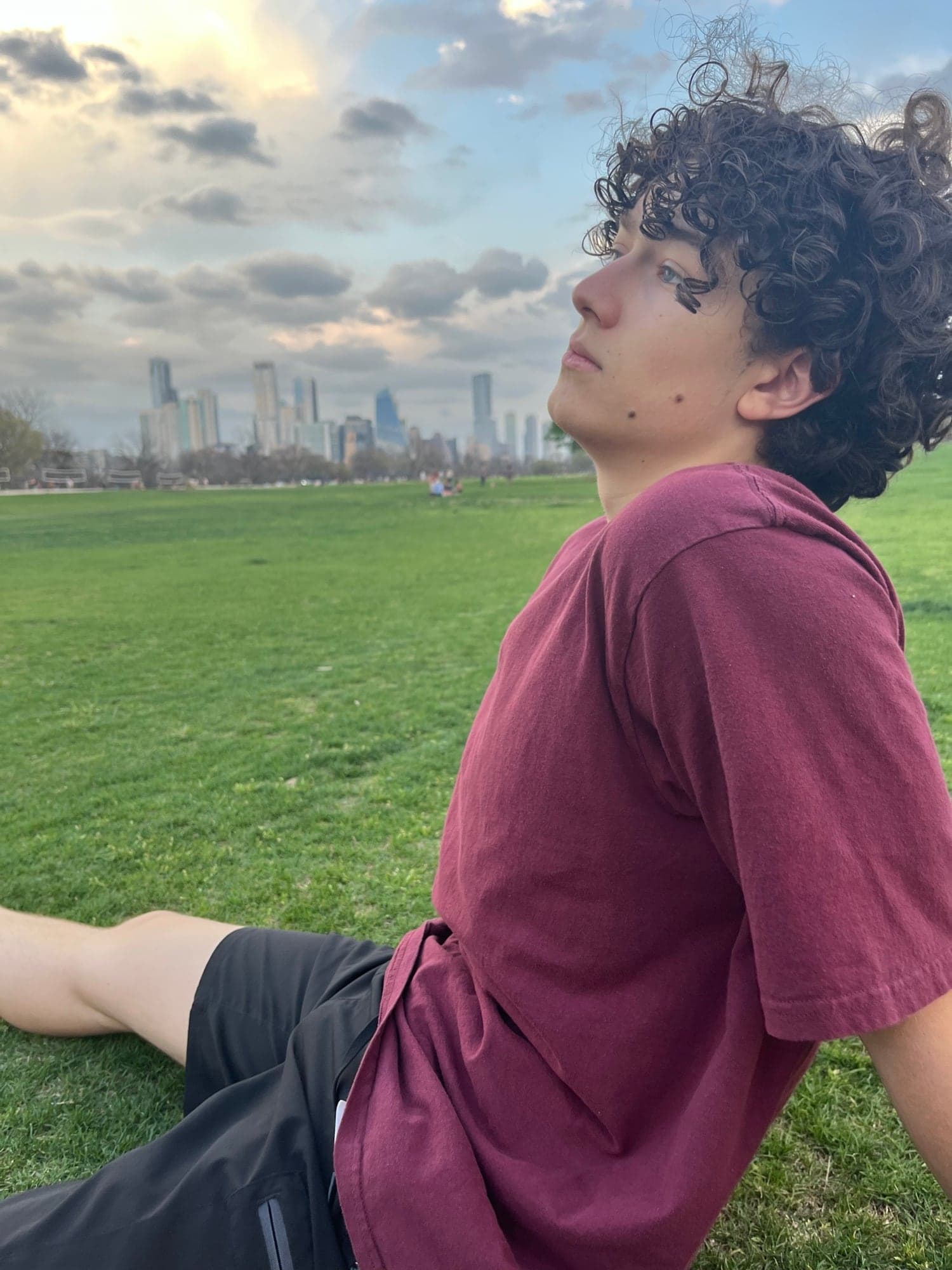 Cruce sitting on the grass at an Austin park in a maroon T-shirt, his face turned up and away from the camera, the downtown skyline and a cloudy sky behind him.